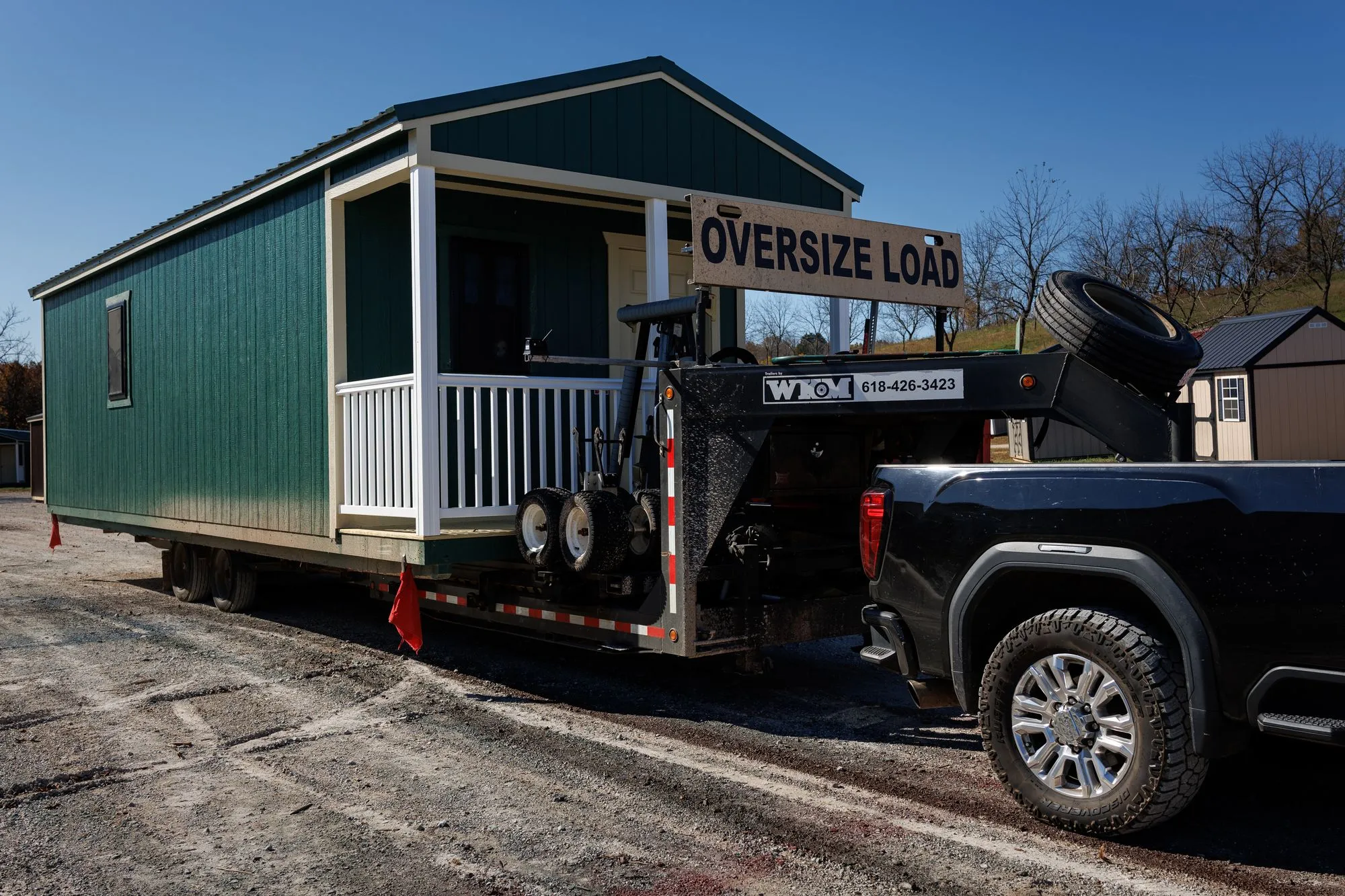A green Hawthorn cabin with a white porch loaded on a delivery trailer behind a pickup truck