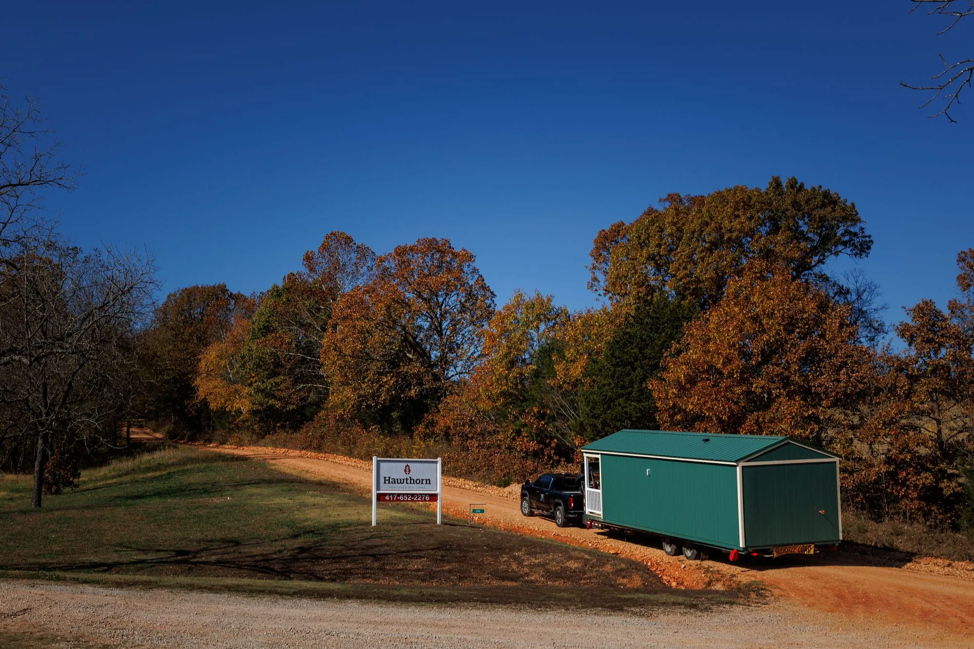 A Hawthorn cabin on its delivery trailer driving out of the lot past the Hawthorn sign, with fall trees behind