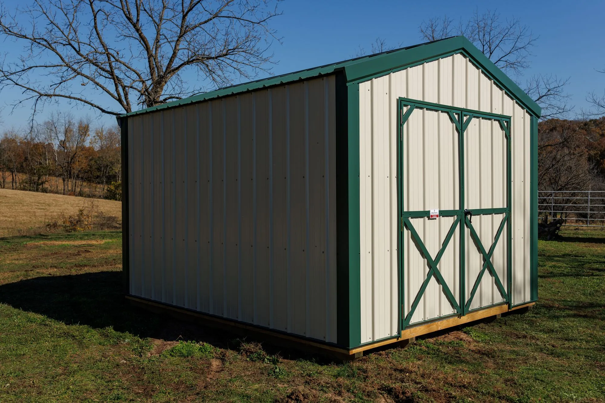 Utility Shed with ivory metal siding with green trim and roof, green X-braced doors, on the Hawthorn lot in Rocky Comfort, Missouri