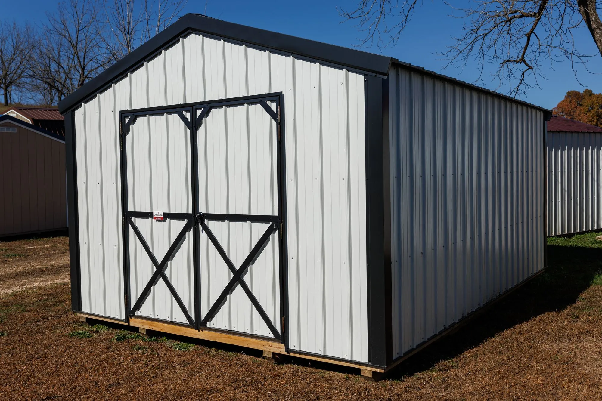 Utility Shed with white metal siding, black trim and roof, black X-braced doors, on the Hawthorn lot in Rocky Comfort, Missouri