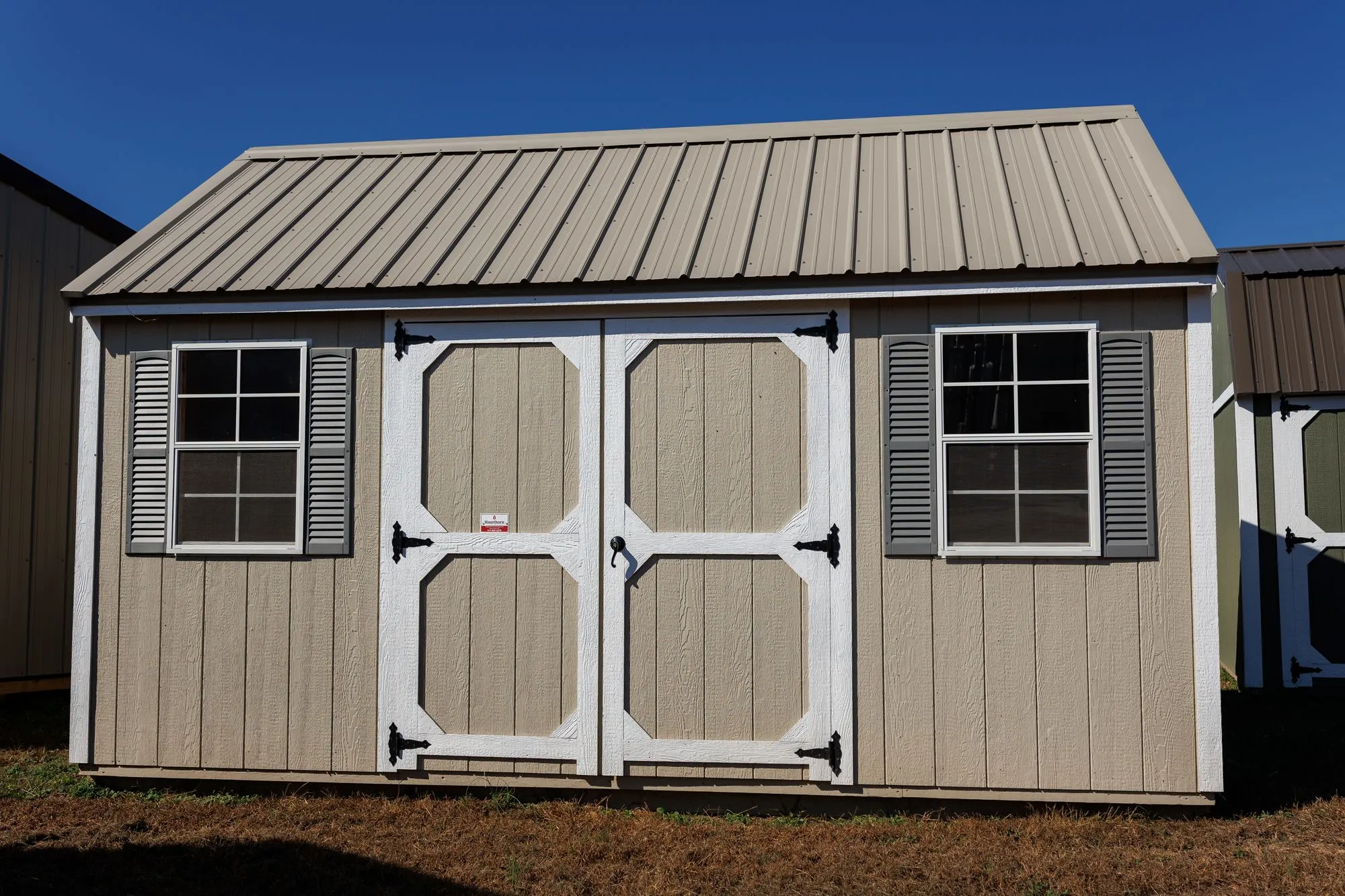 Lofted Garden Shed with tan siding, white trim, clay roof, white octagon-panel doors, two windows, on the Hawthorn lot in Rocky Comfort, Missouri