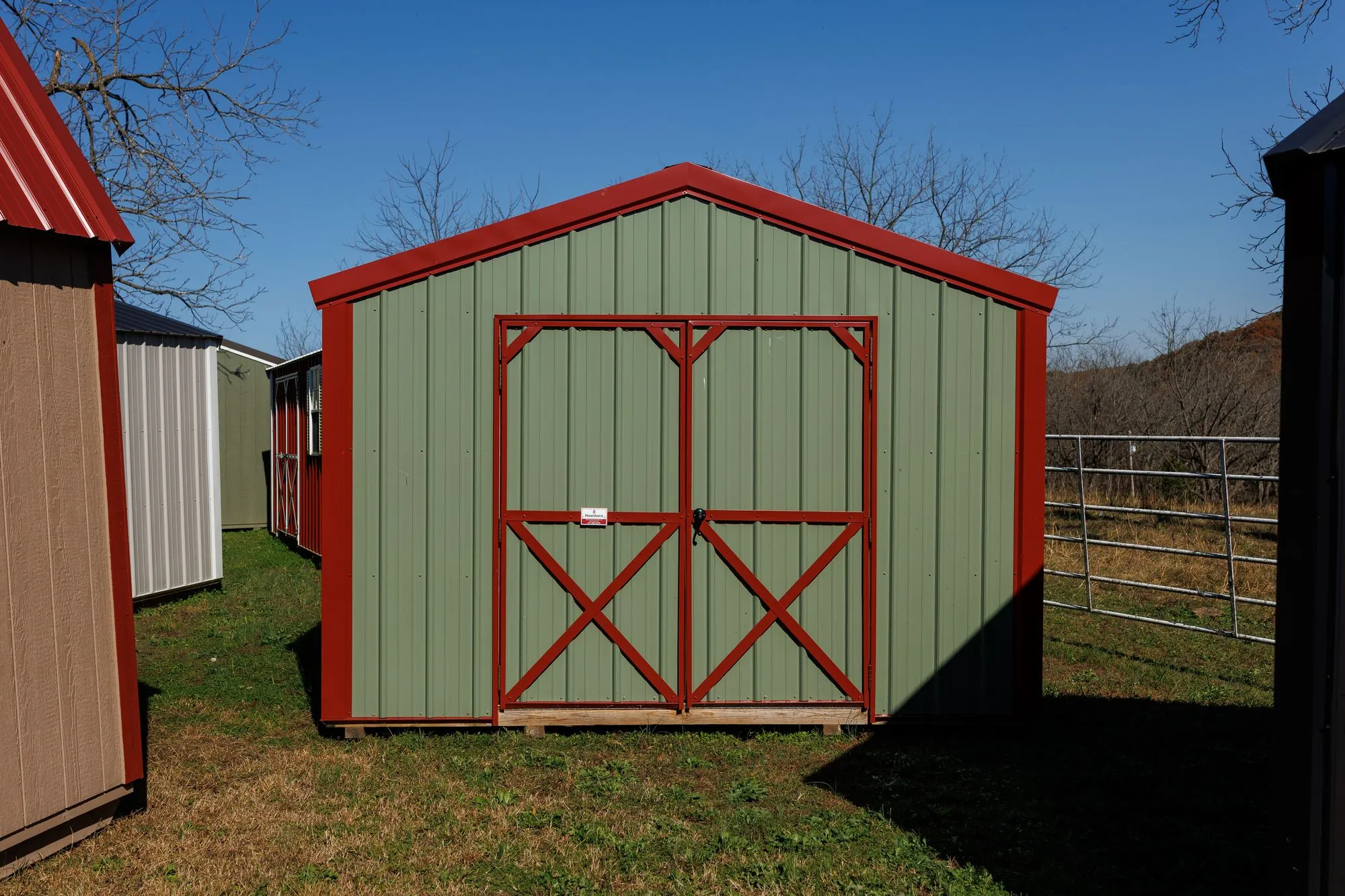 Utility Shed with sage metal siding, red trim and roof, red X-braced doors, on the Hawthorn lot in Rocky Comfort, Missouri